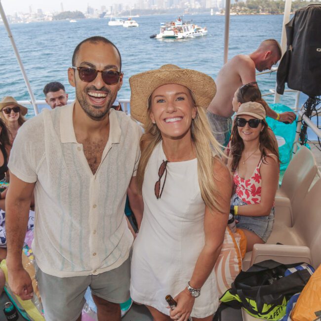 A smiling man and woman in summer clothes and sunglasses pose on a boat with friends. Other people sit and relax in the background, with water and boats visible outside. The mood is festive and casual.