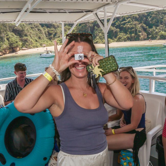 A woman on a boat holds keys and an object to her eye, smiling playfully at the camera. Other people sit and laugh in the background, with clear water and green hills visible outside.