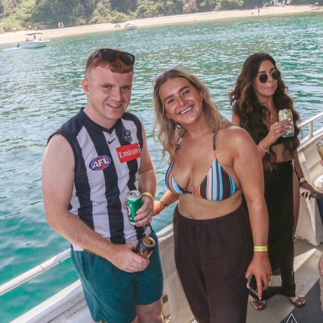 Three people stand on a boat by the water; one man in sportswear holds a drink, and two women, one in a striped bikini top and another in black, smile at the camera on a sunny day. Trees and a beach are visible in the background.