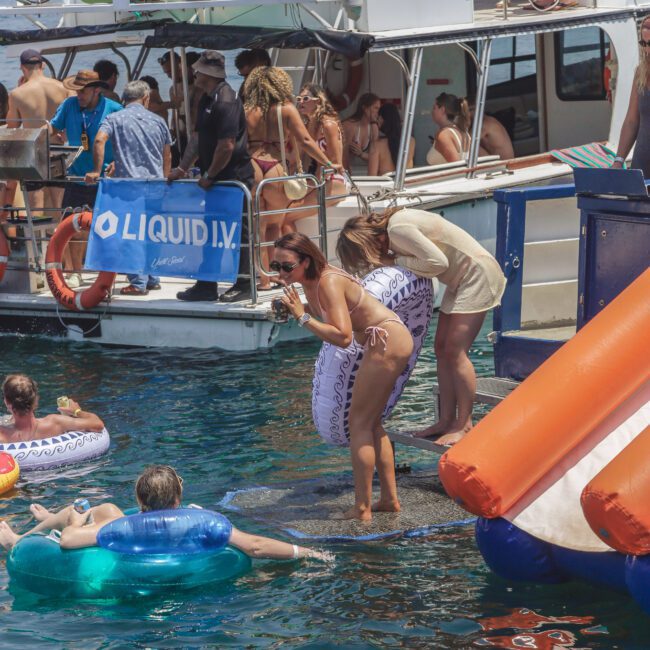 A lively party scene on the water with people on a boat and in the water using inflatables. Two women stand near the edge of the boat, and many are relaxing and socializing under the sun.