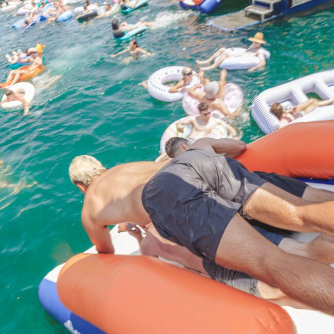 Two men slide down an inflatable slide into a crowded, sunlit lake filled with people relaxing on colorful floaties and pool toys. The scene is lively and festive.