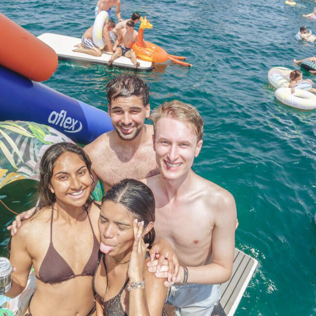 Four young adults in swimsuits smile and pose on a dock by the water, surrounded by boats and inflatable floats, enjoying a sunny day with others at a lively party.
