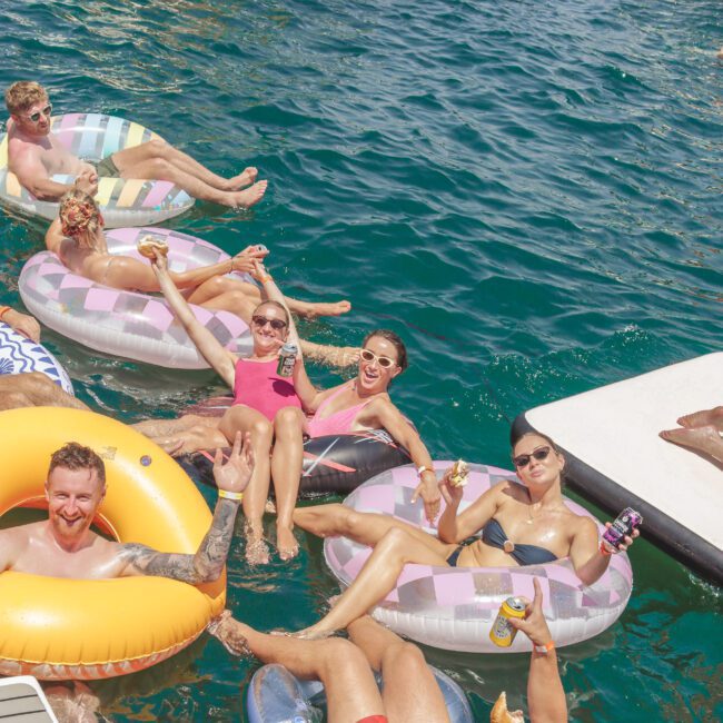 A group of six smiling people relax on colorful inflatable tubes, holding drinks and enjoying sunny weather on the water near a dock, with boats visible in the background.