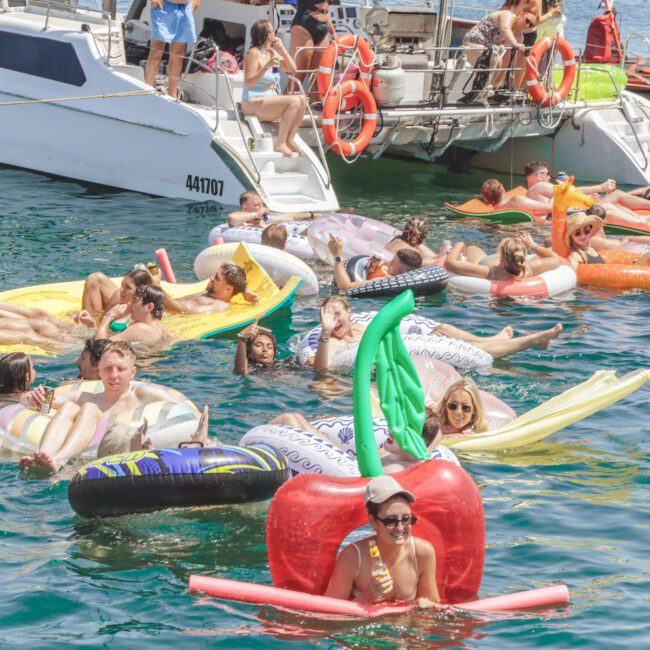A group of people relax on colorful inflatable pool floats in the water near docked boats on a sunny day, enjoying a lively party atmosphere.