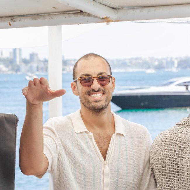A man wearing sunglasses and a light-colored shirt smiles and waves at the camera on a boat. The background shows water, a black yacht, and distant buildings. Another person with a cap stands beside him, facing away.