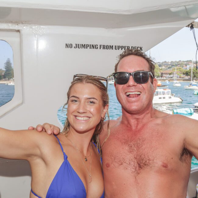 A smiling woman in a blue swimsuit and a shirtless man wearing sunglasses pose for a selfie on a boat, with water, boats, and a shoreline visible in the background. A sign reads "NO JUMPING FROM UPPER DECK.