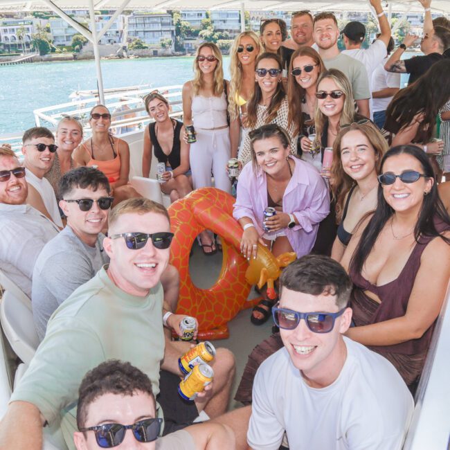 A group of smiling young adults pose for a photo on a boat, some holding drinks. There’s a large inflatable orange float in the center. Water and buildings are visible in the background on a sunny day.