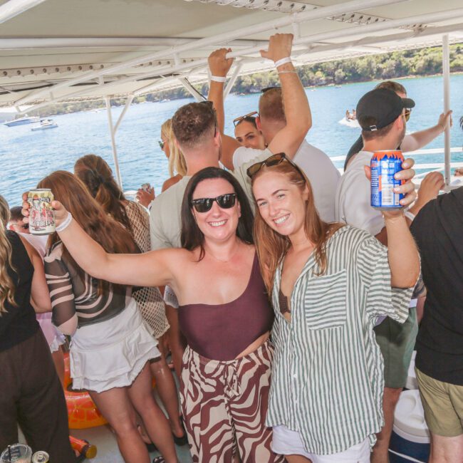 A group of people are smiling, dancing, and raising drinks on a boat. Two women in front pose with cans, enjoying a sunny day on the water. Other guests are socializing in the festive, outdoor atmosphere.
