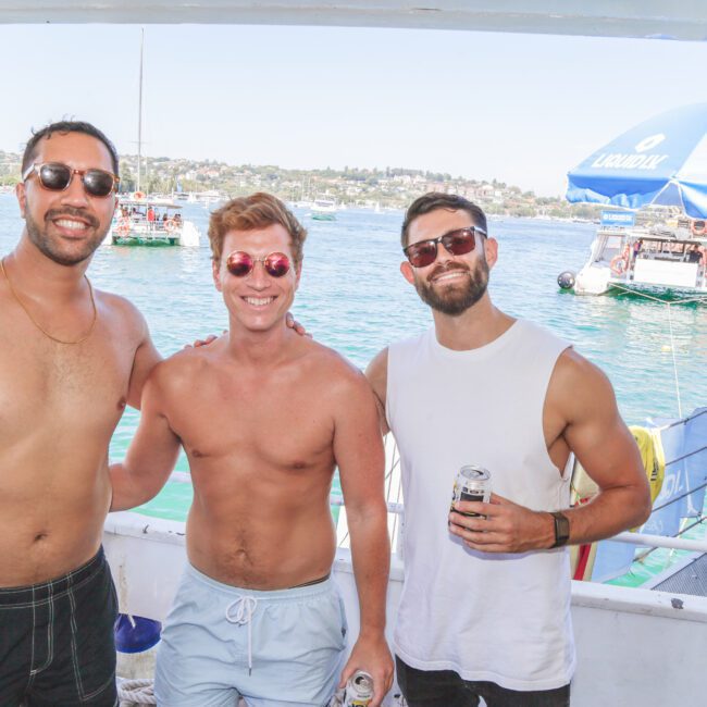 Three men in swimwear and sunglasses smiling and holding drinks on a boat, with water, other boats, and a blue Yacht Social umbrella visible in the sunny background.