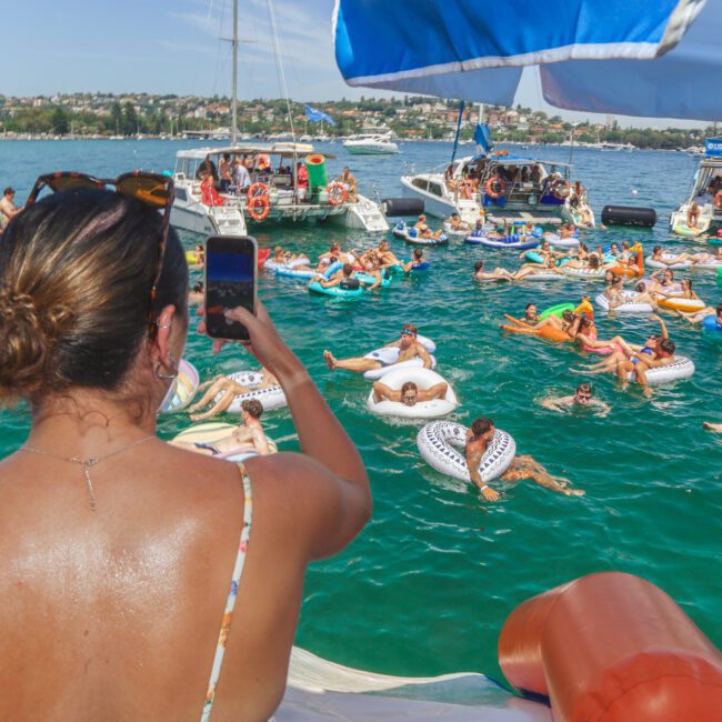 A woman on a boat takes a photo of a lively scene: people relaxing on colorful pool floats in the water, surrounded by boats, under a sunny sky at a social yacht event.