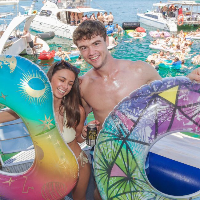 A young woman and man in swimsuits smile on a boat, each holding a colorful inflatable tube. In the background, people relax on boats and floats in the blue water, enjoying a lively summer gathering.