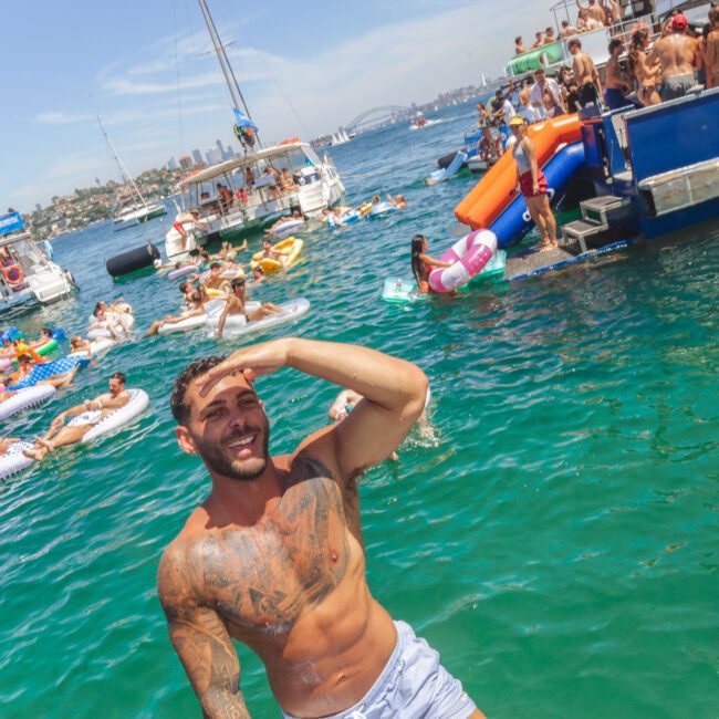 A shirtless man smiles and shades his eyes by the water, surrounded by people on inflatable floats and boats at a lively summer party. The sky is clear and the atmosphere is festive.