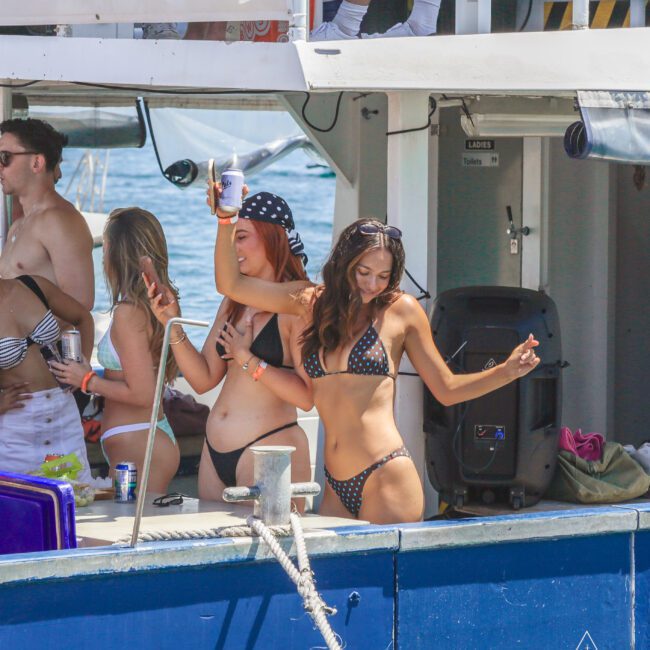 Women in swimsuits dance and pose for selfies on a boat, holding drinks. Other people are in the background, and the scene appears festive and sunny, suggesting a lively party atmosphere on the water.