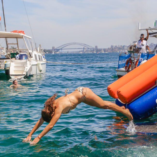 A woman in a swimsuit dives into the ocean from an orange inflatable slide attached to a boat, with other boats, swimmers, and a bridge visible in the background under a clear sky.