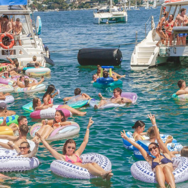 A group of people in colorful swimwear float on inflatable tubes between boats, enjoying a sunny day on the water. Some smile and flash peace signs. A city skyline and houses appear in the background.