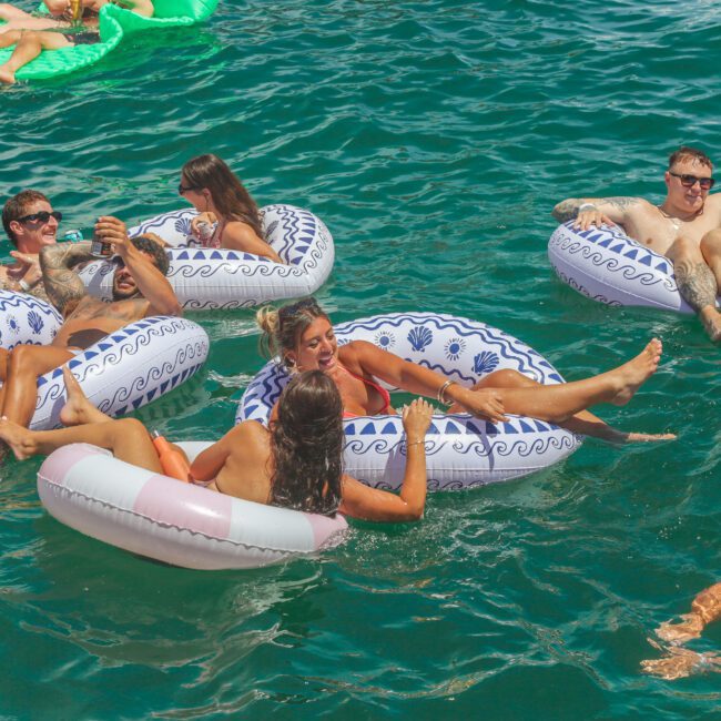 A group of people relax on inflatable pool floats in clear blue water, chatting and enjoying drinks under the sun. Other swimmers are visible in the background.