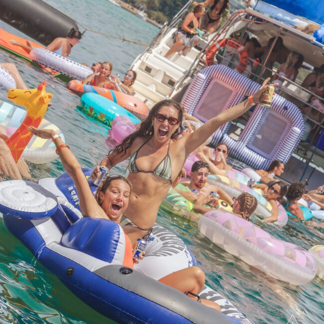 Two women in swimsuits laugh and cheer while floating on inflatable pool toys in the water near a crowded party boat, surrounded by other people and colorful inflatables on a sunny day.
