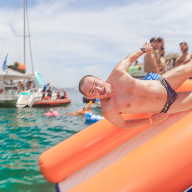 A man in swim trunks slides down an orange and blue inflatable slide into the water, smiling. Several people and boats are in the background, enjoying a sunny day outdoors.
