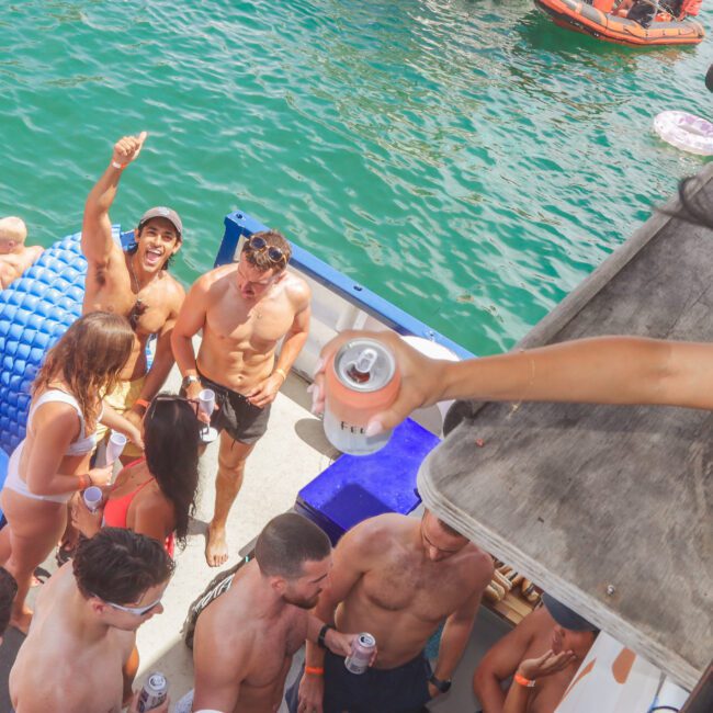 A group of people in swimwear are enjoying a lively boat party on the water. Some are holding drinks, socializing, and smiling. Inflatable floats are nearby, and the sun is shining on the blue water.