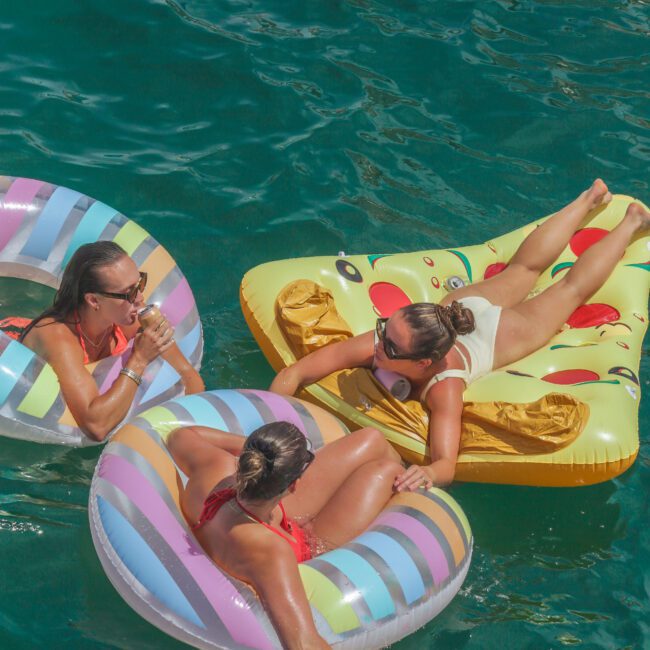 Three women relax on colorful inflatable pool floats in clear blue-green water. Two floats are striped, and one is shaped like a pizza slice. The women are chatting and enjoying the sun.