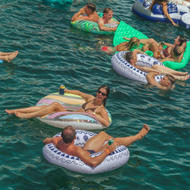 People floating on inflatable pool loungers in a sunny, crowded body of water, relaxing and socializing. Many are holding drinks and wearing swimsuits, enjoying a lively outdoor gathering.