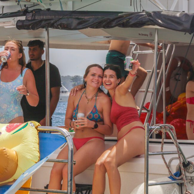 Two women in swimsuits sit smiling and holding drinks on a boat surrounded by friends. Other people in swimwear are standing nearby, with inflatables and water visible in the background. Everyone looks happy and relaxed.
