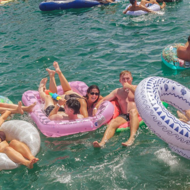 A group of people relax and have fun on inflatable pool floats in a sunny, greenish-blue body of water during a social gathering. Some laugh, chat, and enjoy the lively atmosphere.