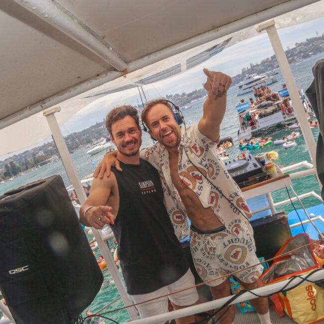 Two smiling men stand behind DJ equipment on a boat, with one wearing headphones and pointing at the camera. The background shows people swimming in the water and boats nearby on a lively, sunny day.