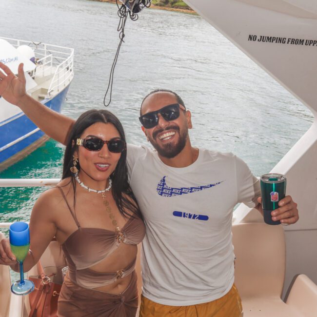 A woman in a brown outfit and a man in a white shirt and yellow shorts smile and pose together on a boat, holding drinks. Water and part of another boat are visible in the background.