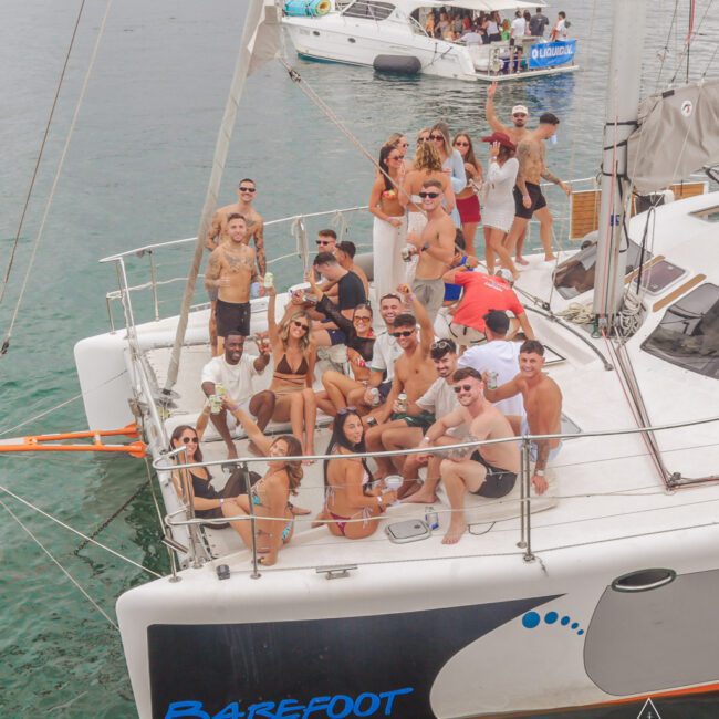 A group of people in swimwear are gathered on the deck of a catamaran named "Barefoot," smiling, posing, and enjoying drinks. Other boats and water can be seen in the background.