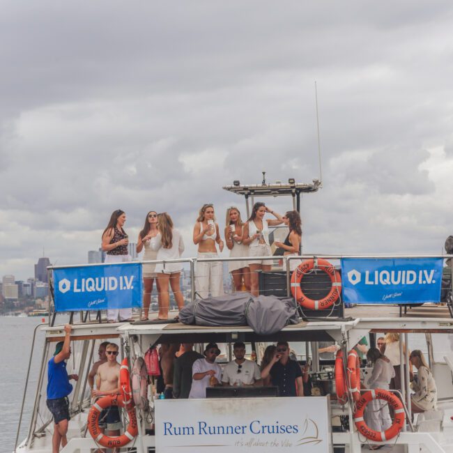 A group of people enjoy a party on a two-level boat labeled "Rum Runner Cruises" with "Liquid I.V." banners, floating on calm water under a cloudy sky, with a city skyline and trees in the background.