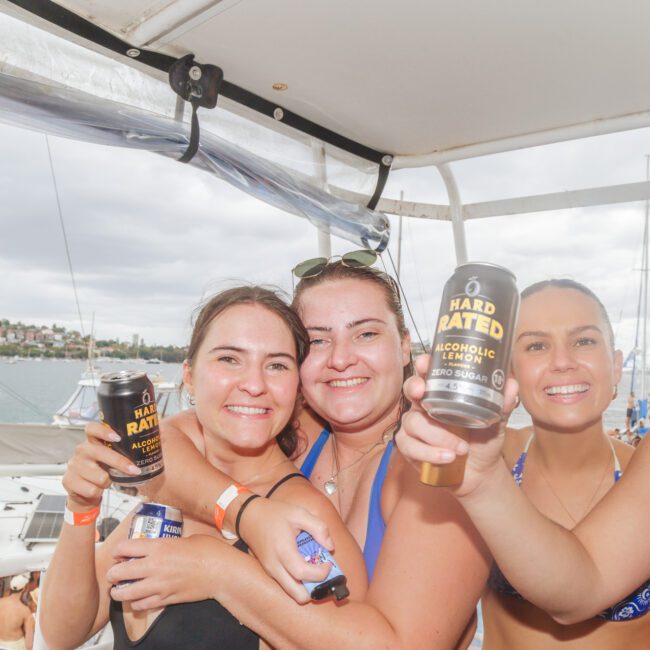Three smiling women in swimsuits hold up cans of Hard Rated alcoholic lemon drink on a boat with other boats and water in the background, enjoying a sunny day at a social event.