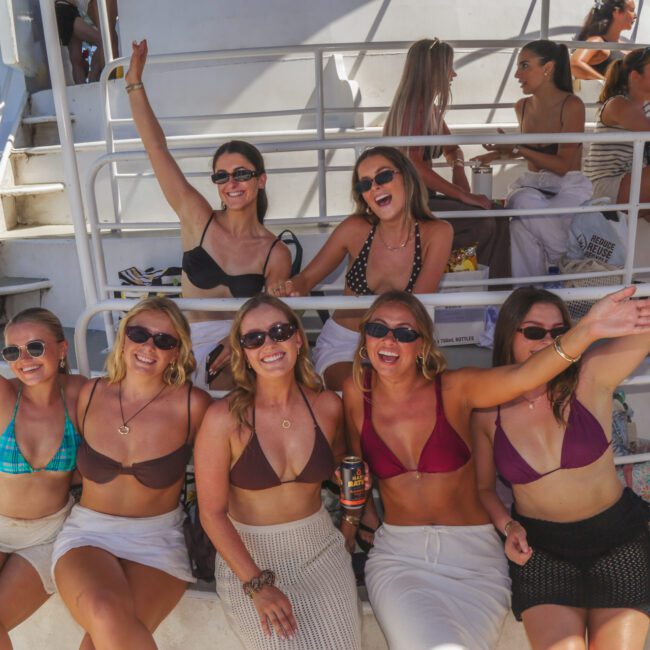 A group of six women in swimsuits and sunglasses sit on a boat, smiling and raising their arms, with more people and blue sky in the background. The atmosphere is cheerful and sunny.
