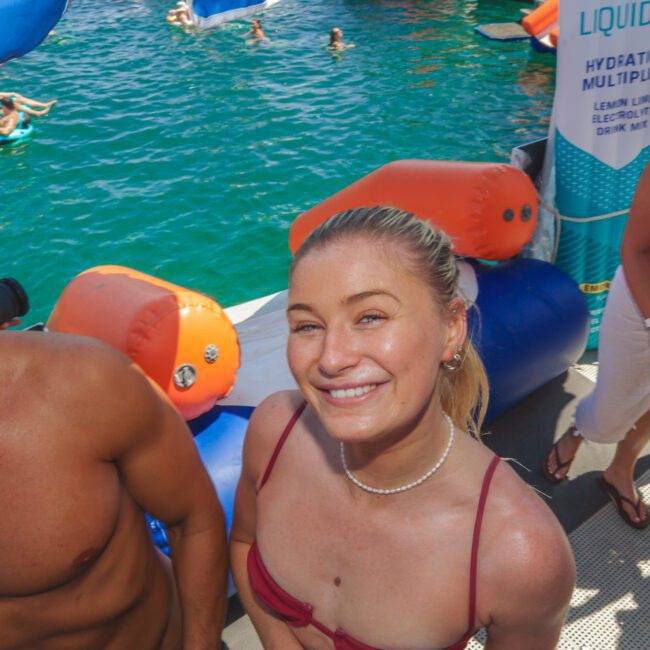 A smiling woman in a pink bikini stands on a boat deck surrounded by people, with blue water and inflatables in the background on a sunny day.