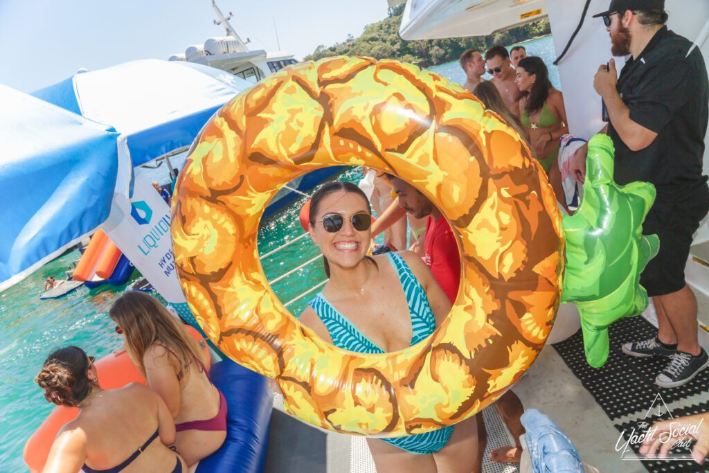 A woman in sunglasses and a striped swimsuit smiles while holding a pineapple-shaped float on a boat with people in swimsuits, inflatable toys, and blue water in the background.