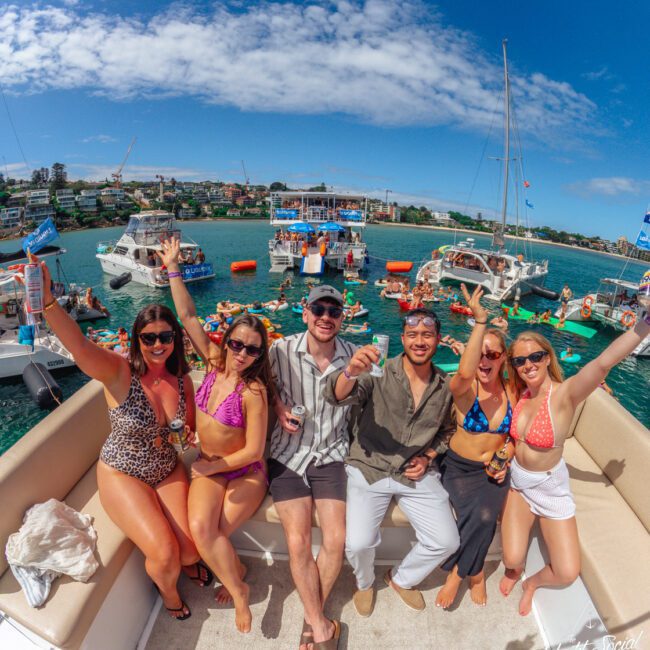 A group of six smiling friends in swimsuits and sunglasses sit on a boat, raising drinks and celebrating. Other boats and people enjoying the water are visible in the sunny background.