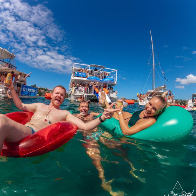 Three people smile and pose on colorful floaties in clear blue water, with boats and a lively group of people in the background under a sunny sky.