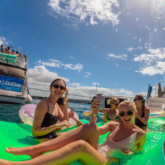 Four women in swimsuits relax on a large green inflatable float, smiling and holding drinks, with boats and blue sky in the background during a sunny day on the water.