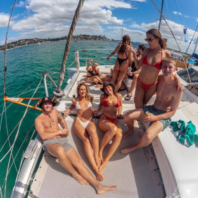 A group of young people in swimsuits relax and pose on the deck of a sailboat under a sunny sky, with turquoise water and a city skyline in the background. Some are holding drinks and smiling at the camera.
