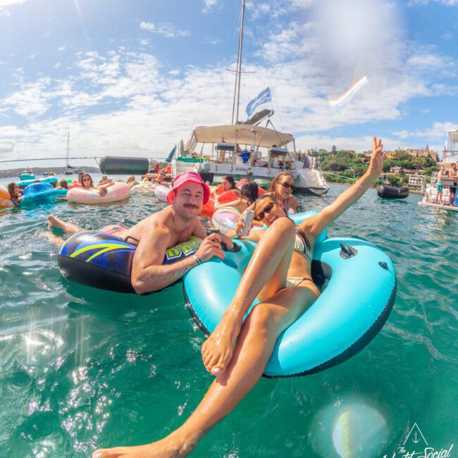 People relaxing on colorful floaties in turquoise water near a yacht. The sky is blue with some clouds, and a woman makes a peace sign while others smile and enjoy the sunny day. The scene is lively and joyful.