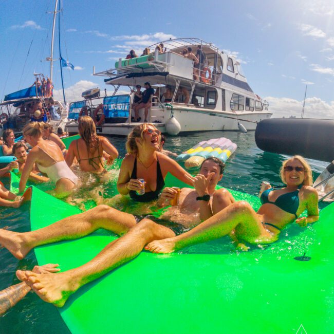 A group of people in swimsuits lounge and laugh on a bright green float in the water near yachts, enjoying a sunny day with drinks and friends at a yacht party.