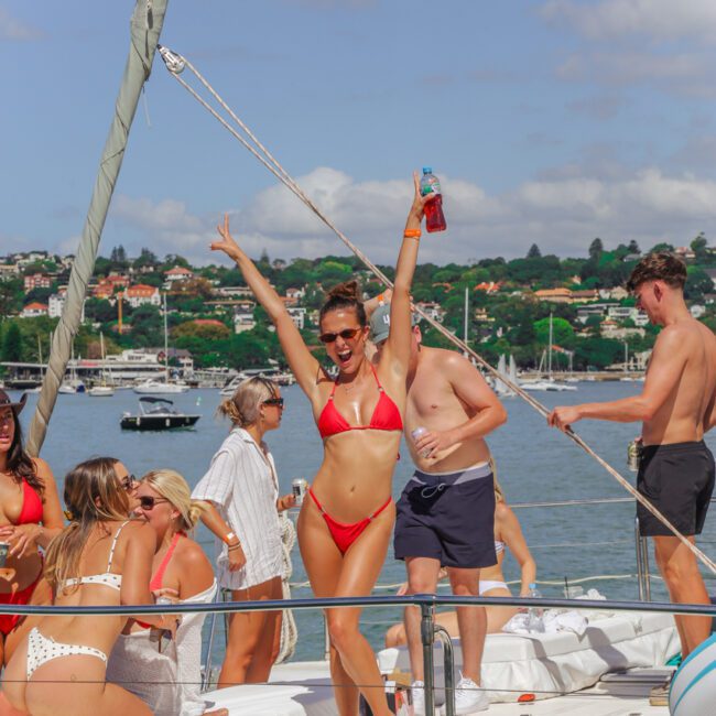 A woman in a red bikini poses with arms up, smiling on a yacht with friends. Others relax and talk nearby. There are boats and greenery in the background under a partly cloudy sky.