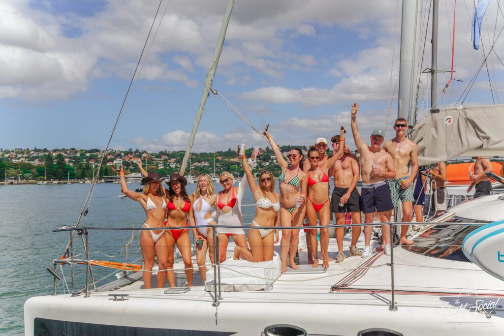 A group of people in swimsuits pose and smile on the deck of a sailboat anchored near the shore, raising their arms and enjoying a sunny day with water and a coastal town in the background.