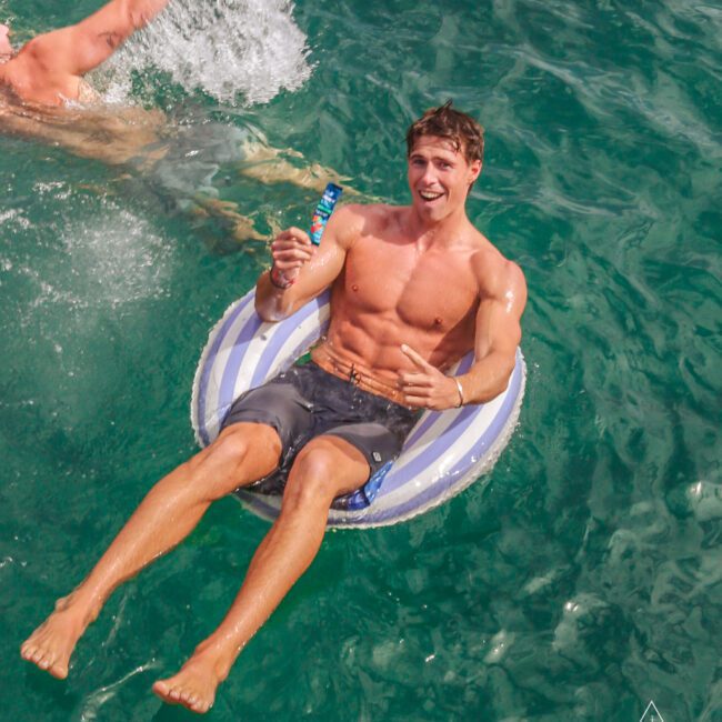 A smiling man lounges on a striped inflatable ring in clear blue water, holding a popsicle. Another person is swimming nearby, splashing water. The scene is bright and sunny, evoking a fun, summer atmosphere.