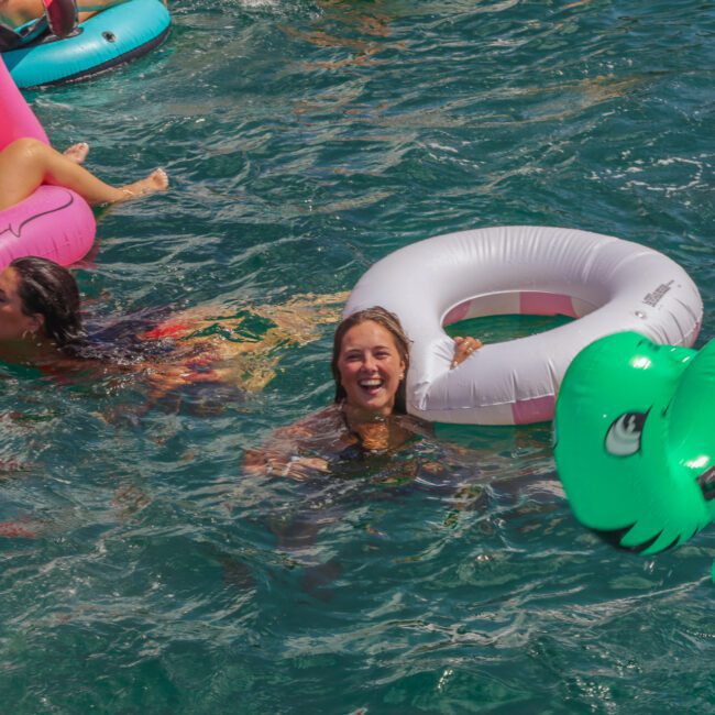 A group of people enjoy swimming and relaxing on colorful inflatable pool floats in clear blue water. One woman smiles at the camera while others lounge nearby. The scene is lively and bright.