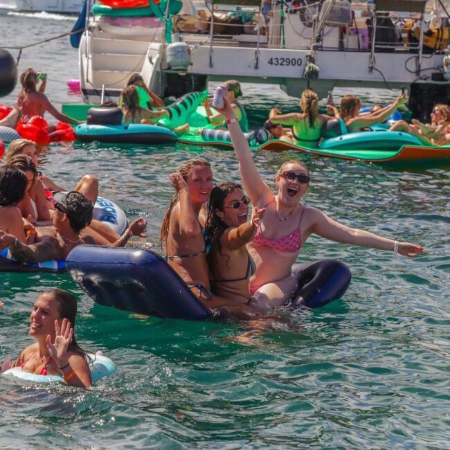 A group of people in swimsuits enjoy a lively pool party on colorful inflatables in the water, smiling, waving, and having fun near docked boats on a sunny day.
