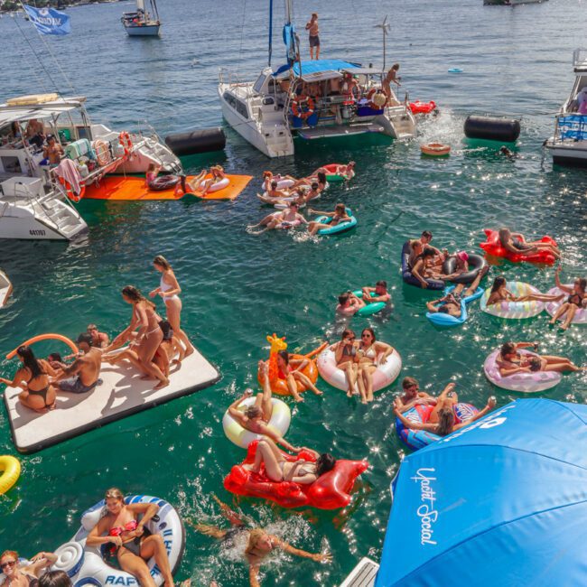 People relax and socialize on colorful inflatables and boats in a sunny harbor, with clear blue water, yachts, and city skyline in the background. A large blue umbrella and floaties are visible in the foreground.