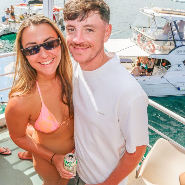 A smiling woman in a pink bikini and sunglasses stands next to a man in a white T-shirt on a boat, with other people and another boat visible on the water in the background.