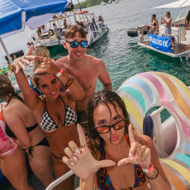 A group of young adults in swimsuits have fun on a boat, posing and smiling for the camera. There are inflatables, other boats on the water, and a sunny sky in the background.