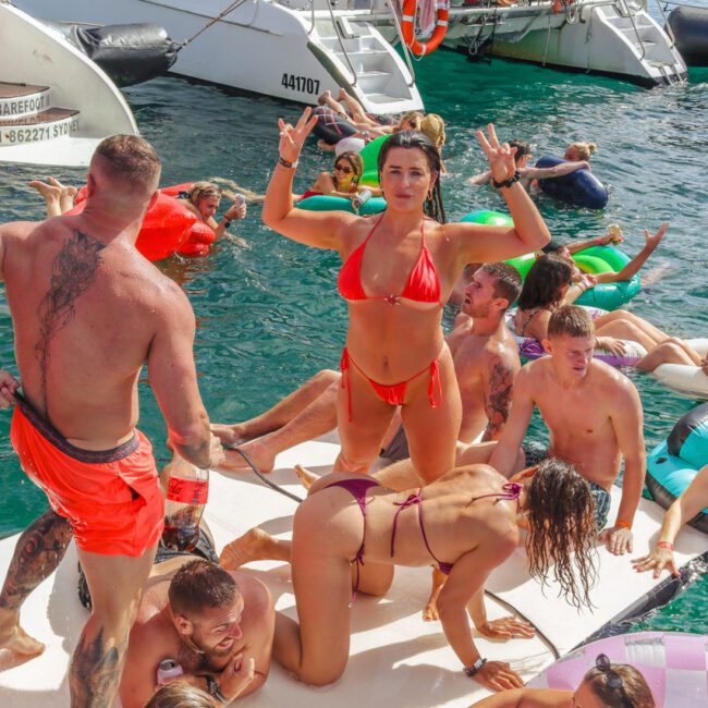 A group of people in swimsuits enjoy a lively party on the water, some on a floating mat and others in the water. A woman in a red bikini poses with hand signs while boats are seen in the background.